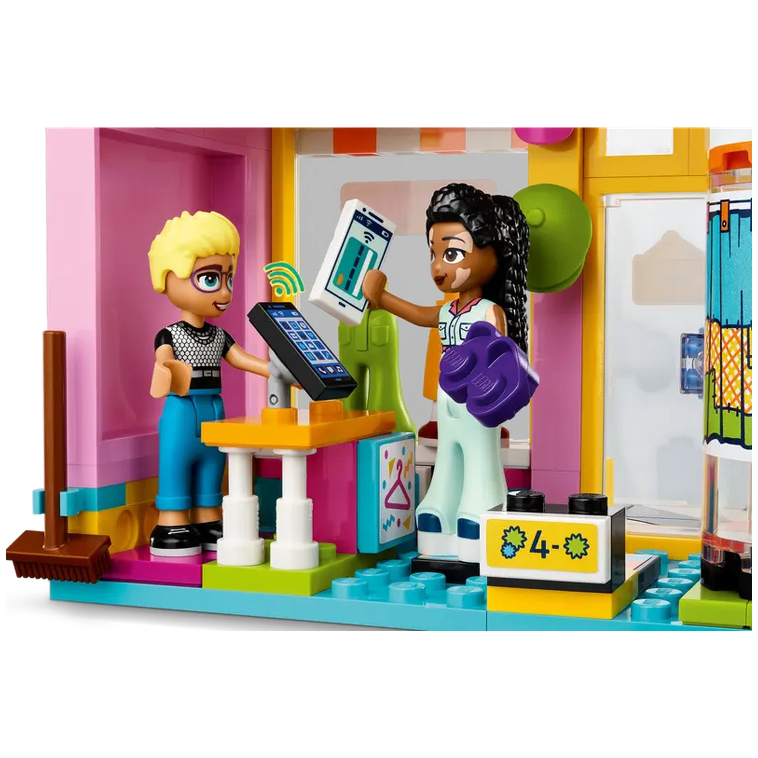 Two LEGO Friends characters interact in a colorful Vintage Fashion Store. One girl, with a blonde hairstyle, stands at a checkout counter featuring a tablet, while the other girl, holding purple accessories, displays a phone. The vibrant playset includes clothing items in the background, encouraging imaginative role play and creativity.