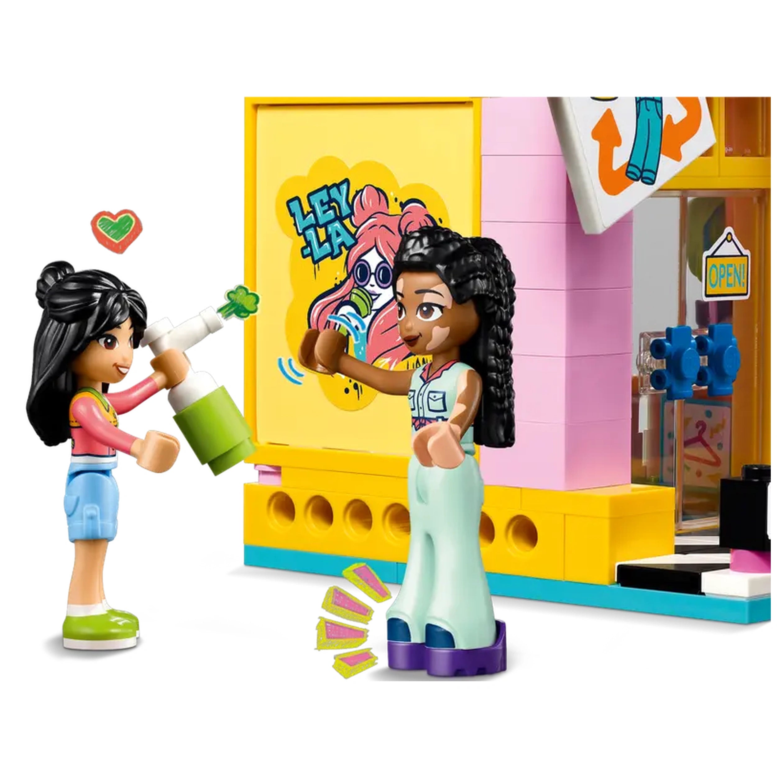 Two LEGO Friends characters interact in front of a colorful Vintage Fashion Store. One character joyfully sprays green paint while the other poses with enthusiasm. The store features a vibrant mural with a pink-haired graphic and an "OPEN" sign.
