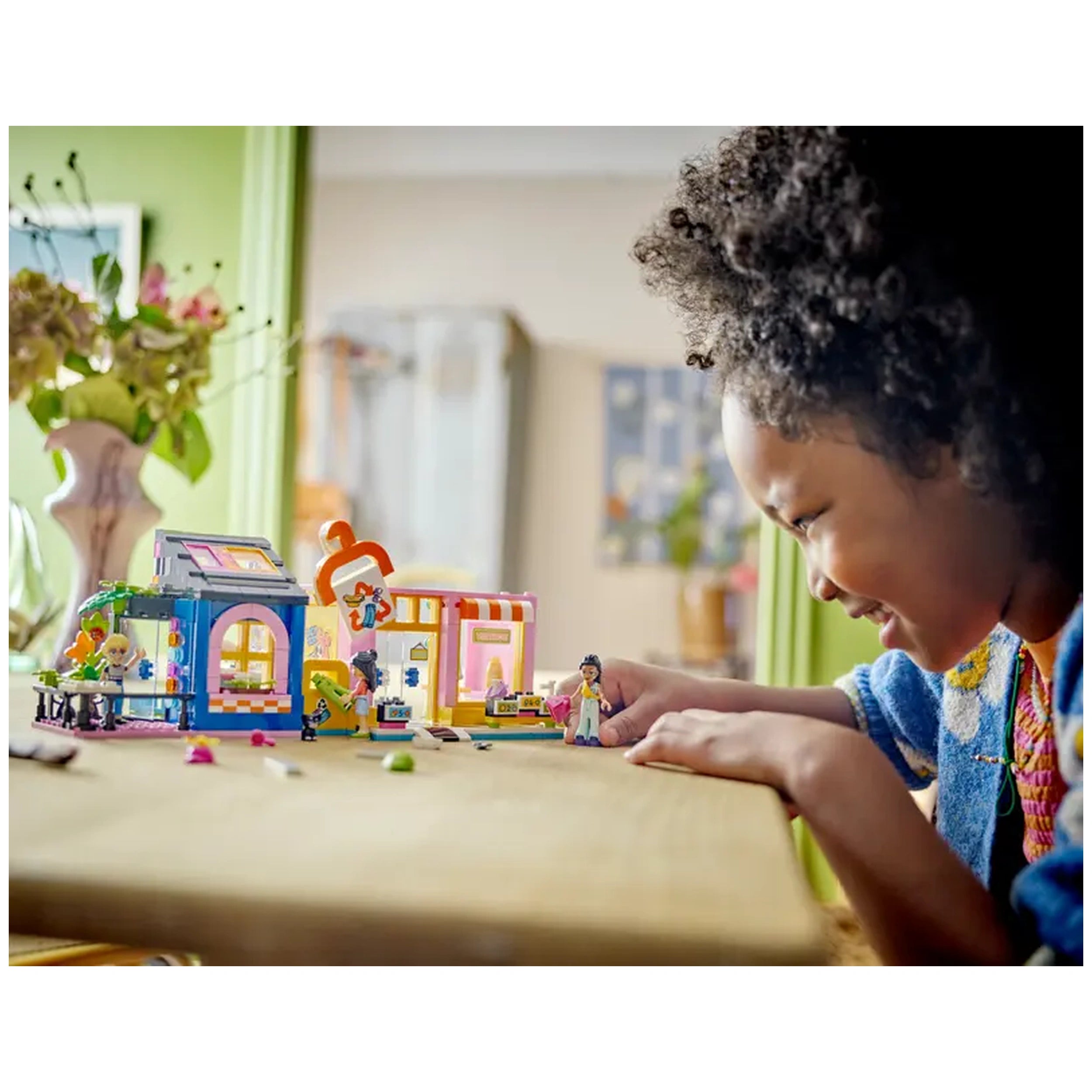A young girl smiles while playing with the colorful LEGO® Friends Vintage Fashion Store set, featuring a blue and pink shopfront, LEGO characters, and various accessories on a wooden table.