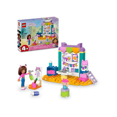 Gabby mini-doll figure stands beside an art station with colorful crafting supplies, a ladder, and a decorated wall, featuring a vibrant playset.
