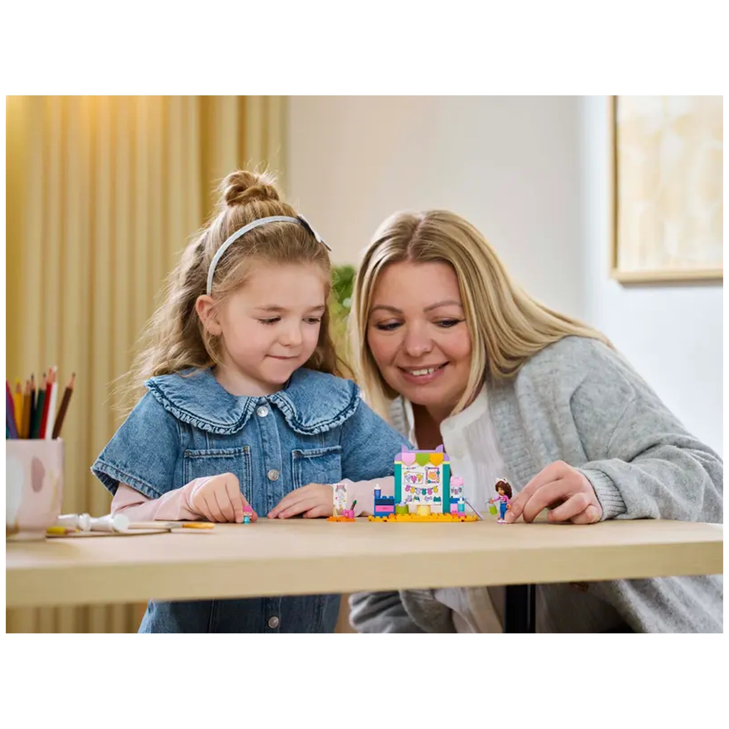 A young girl in a denim dress and a woman with long blonde hair smile while engaging with the LEGO® Gabby's Dollhouse Crafting with Baby Box set on a wooden table, surrounded by colorful crafting tools and art supplies.
