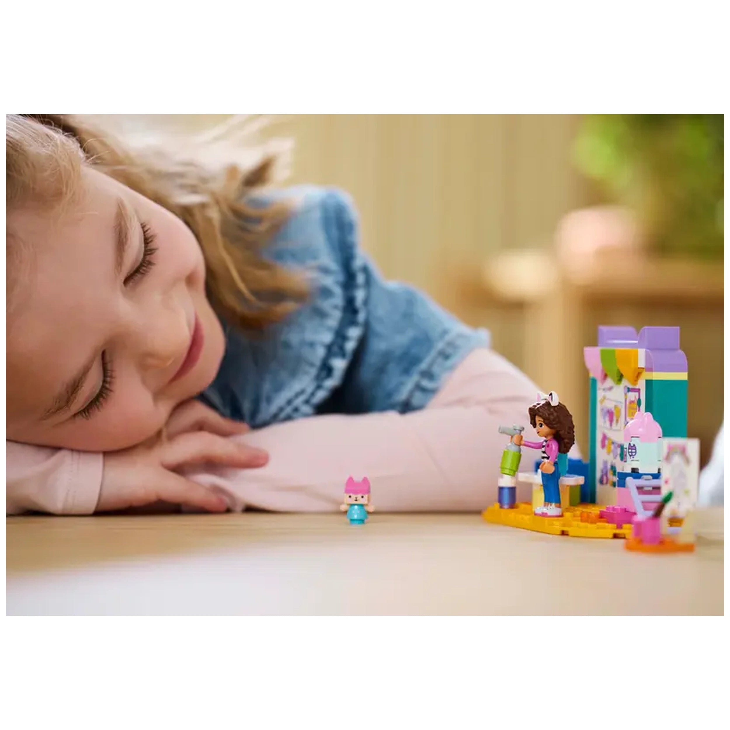 Smiling girl in a denim jacket enjoys playing with the LEGO® Gabby’s Dollhouse Crafting with Baby Box set, featuring colorful characters and crafting elements on a wooden table.