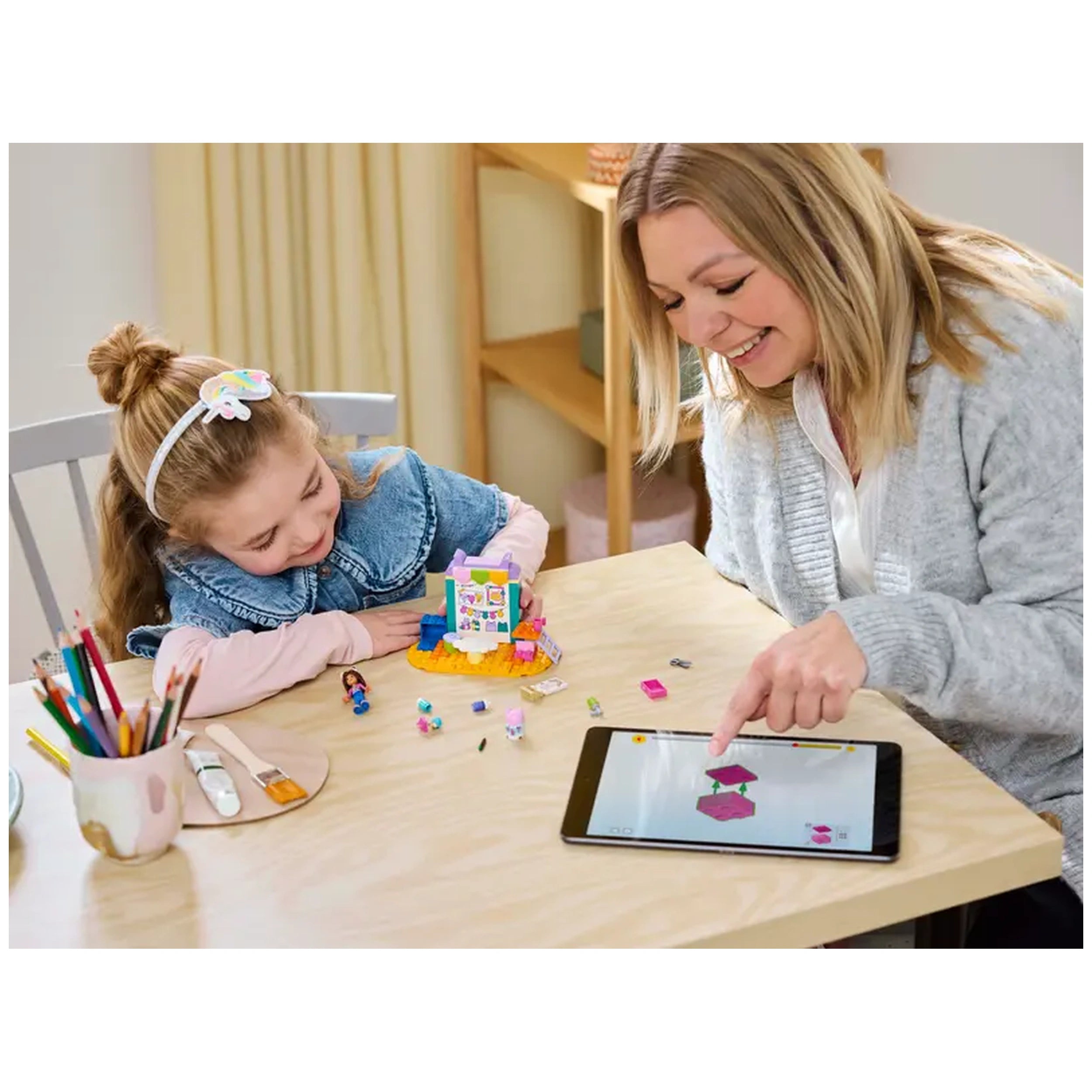 A smiling woman and a young girl interact at a wooden table, exploring the LEGO® Gabby's Dollhouse Crafting with Baby Box set. The girl, wearing a denim jacket and a colorful hair accessory, looks excited as they examine the colorful pieces, including a mini-doll and various crafting supplies. The woman gestures toward a tablet displaying vibrant graphics, showcasing the engaging activities of the set. Colored pencils are neatly arranged nearby, emphasizing a creative atmosphere.