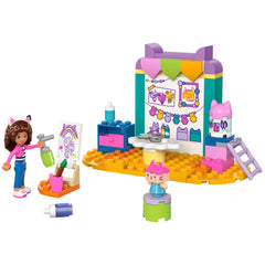 Gabby, a mini-doll figure with curly hair in a pink and white outfit, holds a spray can next to a colorful craft station. The set features a drawing easel with a rainbow picture, crafty supplies, and a Baby Box figure wearing a pink hat, all set in a vibrant play area with blocks in shades of orange, turquoise, and purple.