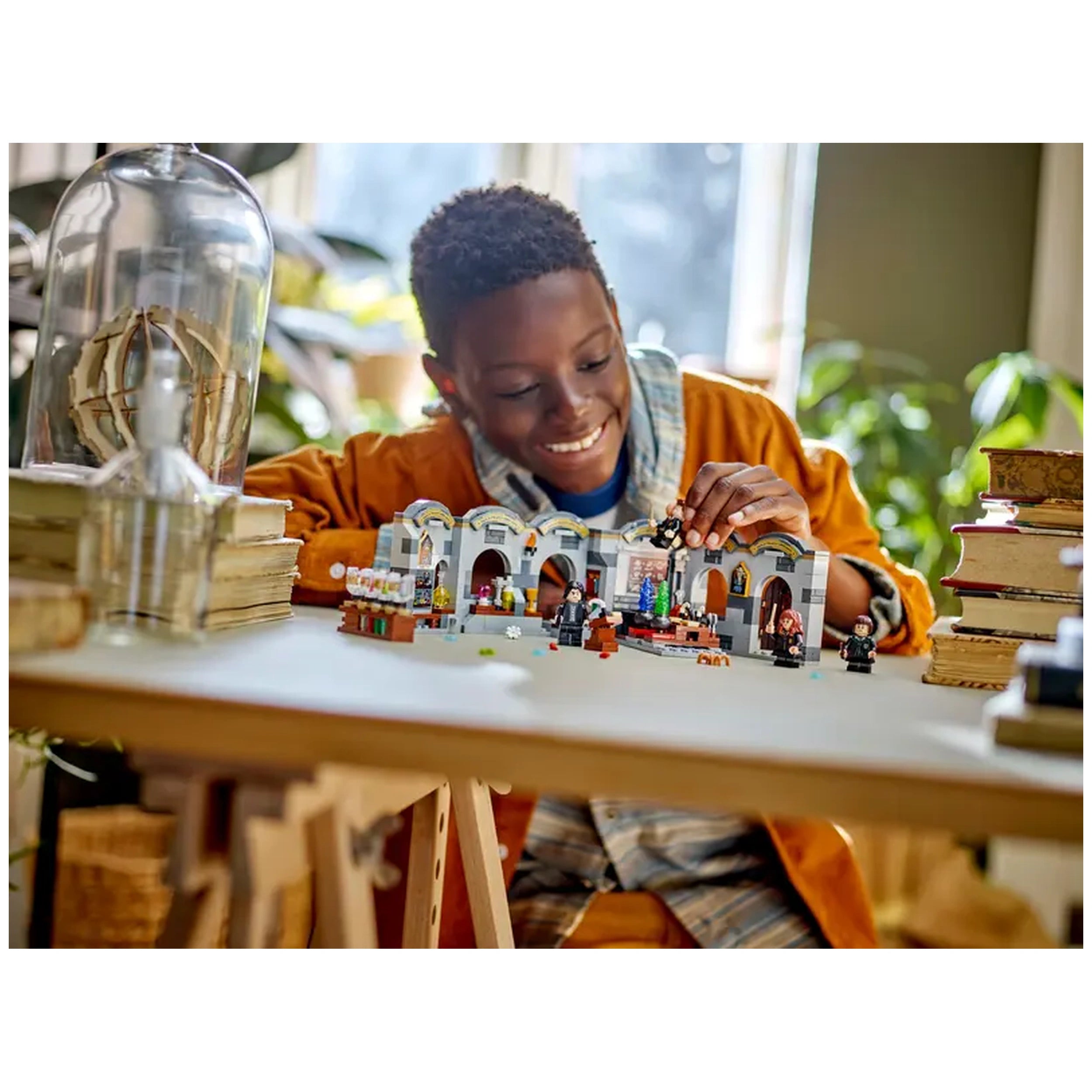 A joyful child wearing an orange jacket builds the LEGO® Harry Potter™ Hogwarts Castle: Potions Class set, featuring detailed classroom elements and minifigures, on a table surrounded by books and plants.