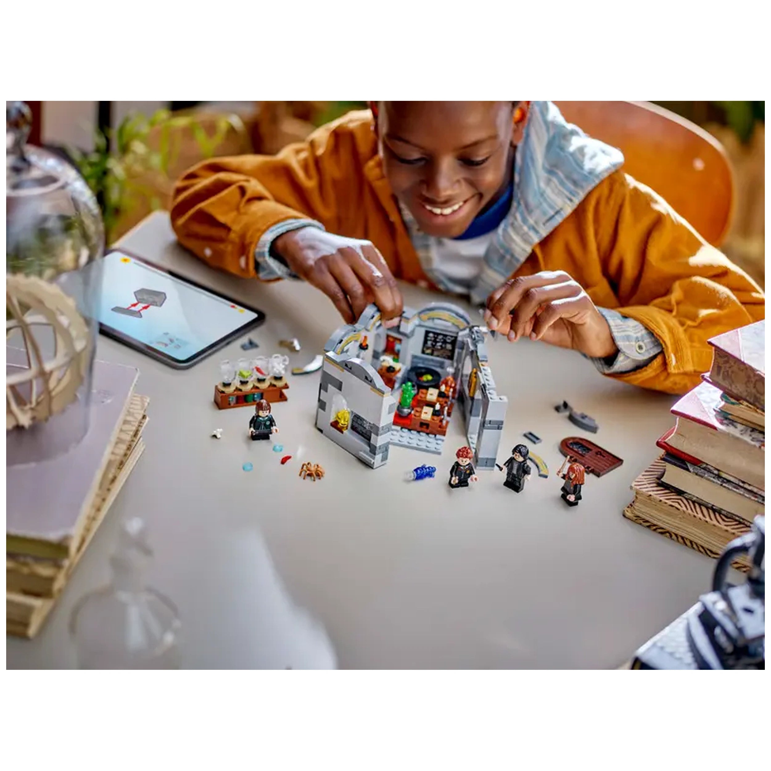 A joyful child builds the LEGO® Harry Potter™ Hogwarts Castle: Potions Class set, featuring colorful minifigures, ingredients, and classroom details on a table with an open instruction tablet.