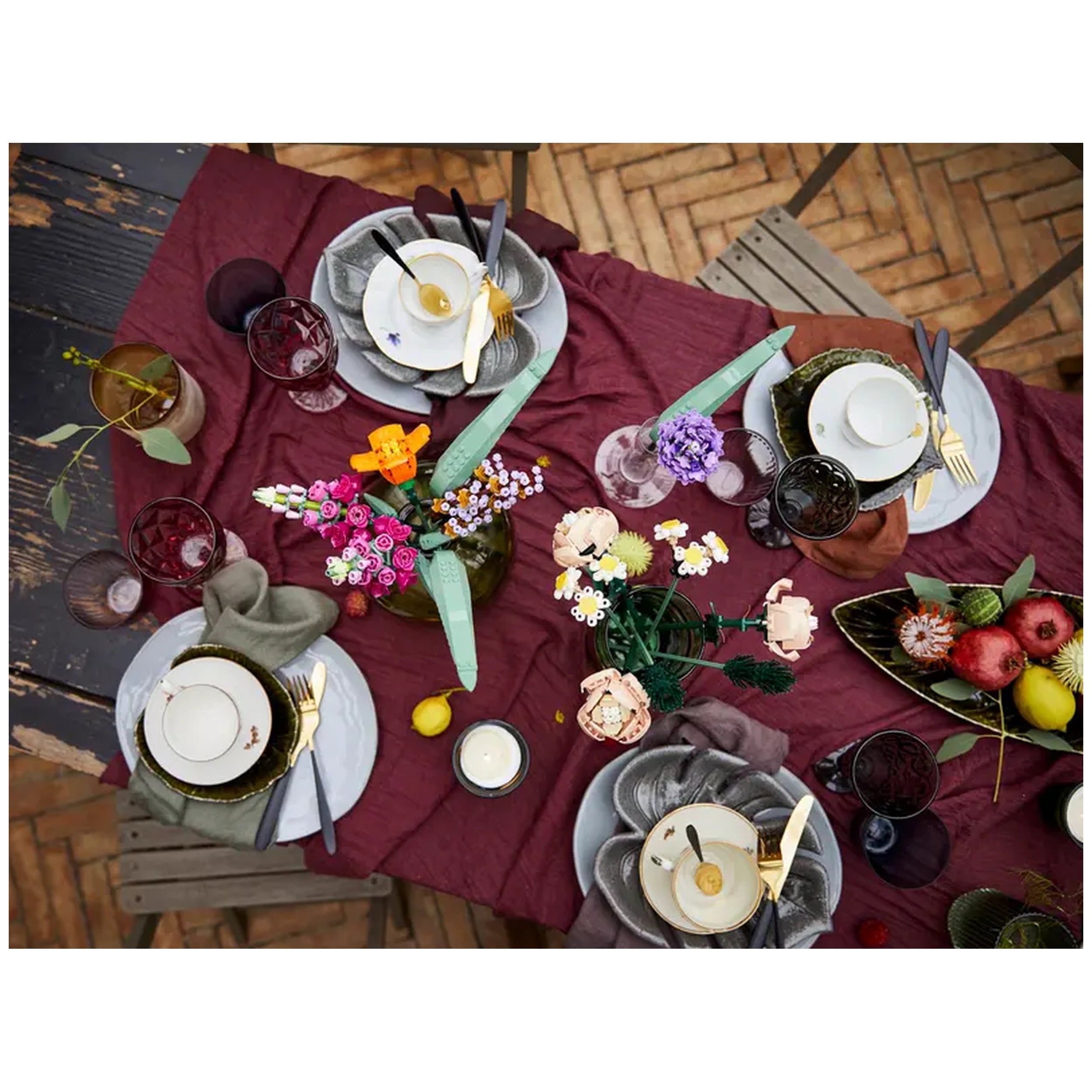 A beautifully arranged table is set with vibrant LEGO Flower Bouquets, elegant dishware, and assorted colorful fruits, featuring a rich burgundy tablecloth and assorted glassware.