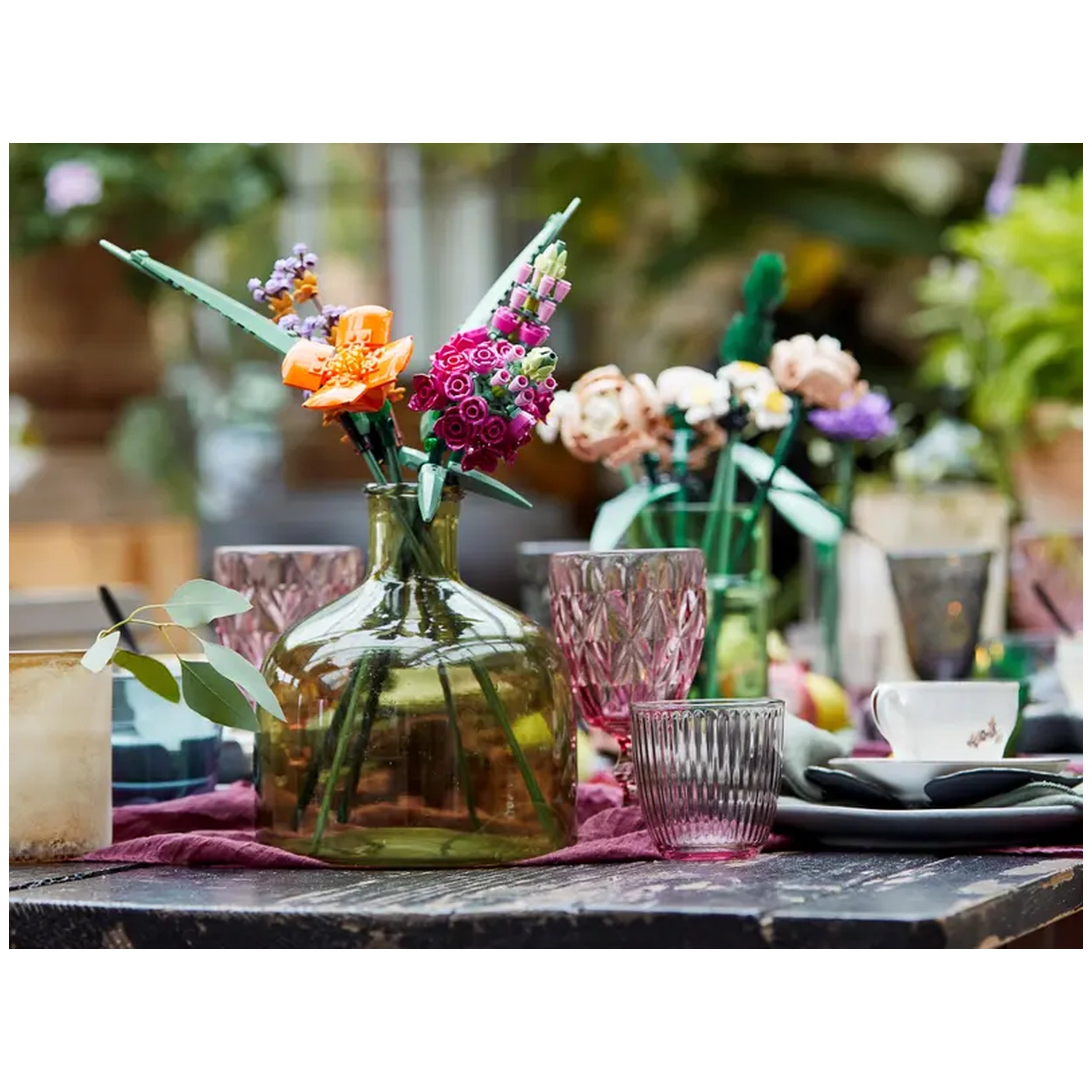 A glass vase filled with colorful LEGO flower creations, including orange blooms and purple roses, sitting on a rustic table with various elegant glassware and decor elements.