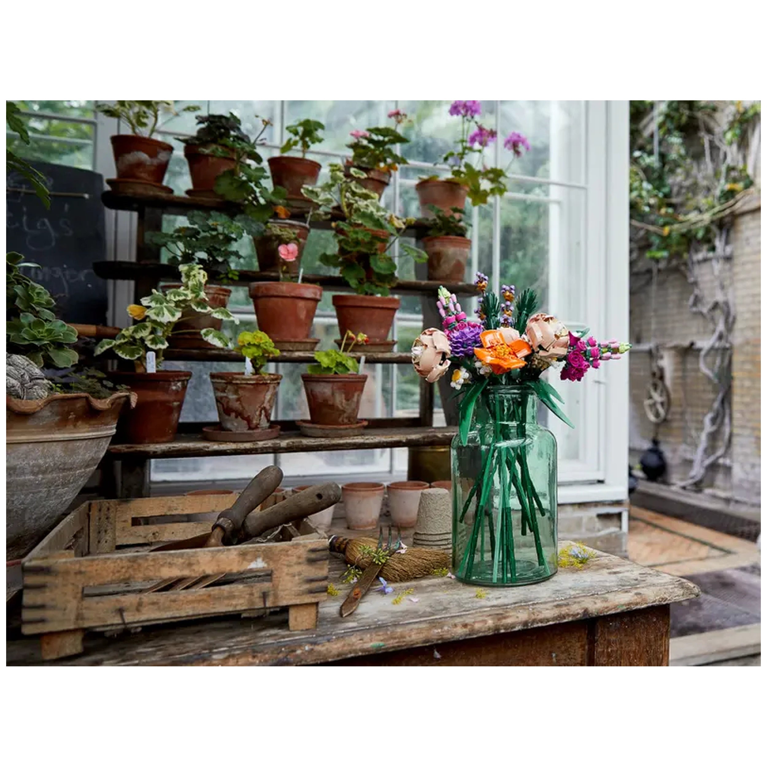A vibrant bouquet made from LEGO flowers, including roses, snapdragons, and daisies, arranged in a green glass vase on a wooden table, surrounded by potted plants.