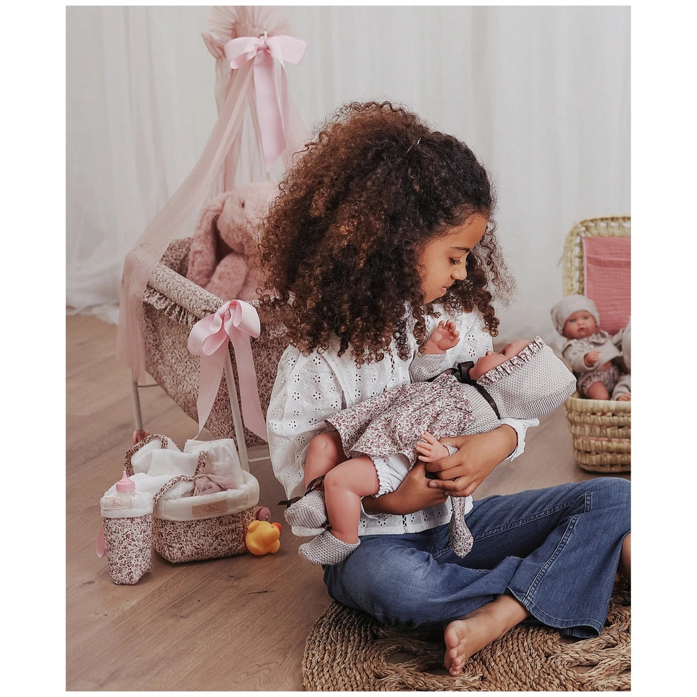 Child playing with Asi Dolls Martina Collection basket, holding a doll beside a pink crib and accessories.