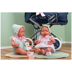Two Asi Dolls in gingham and lace outfits, sitting on round mats with a toy stroller in the background.