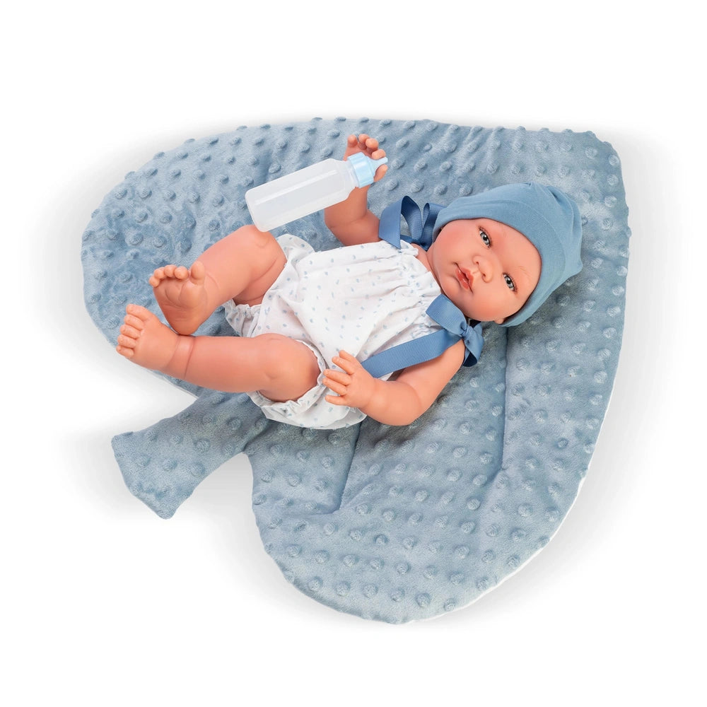 Pablo doll in a romper, lying on a textured blue pillow, holding a bottle, wearing a blue hat.
