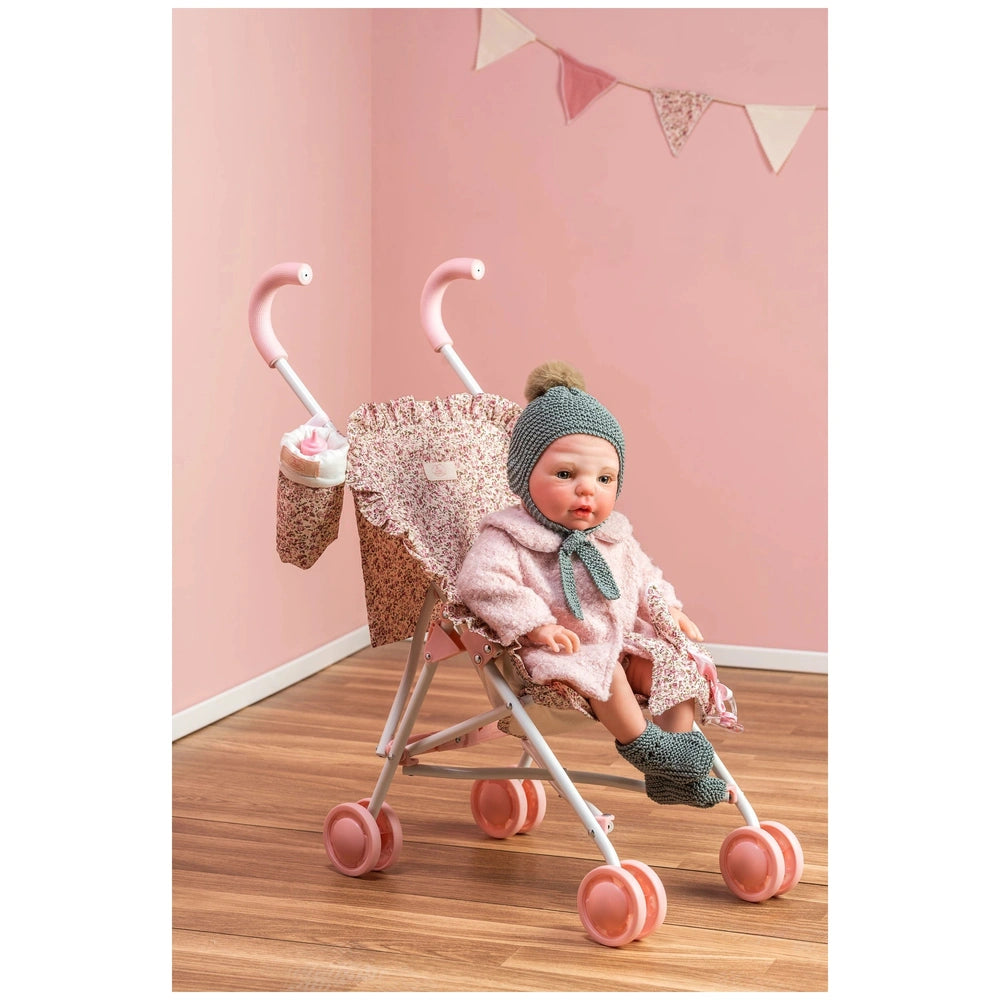 Doll in pink floral stroller with safety straps, pink wheels, and matching bag against a pastel pink wall with bunting.