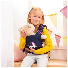 BAYER DESIGN doll carrier in unicorn blue and pink, held by a young girl in a yellow sweater and colorful bunting backdrop.