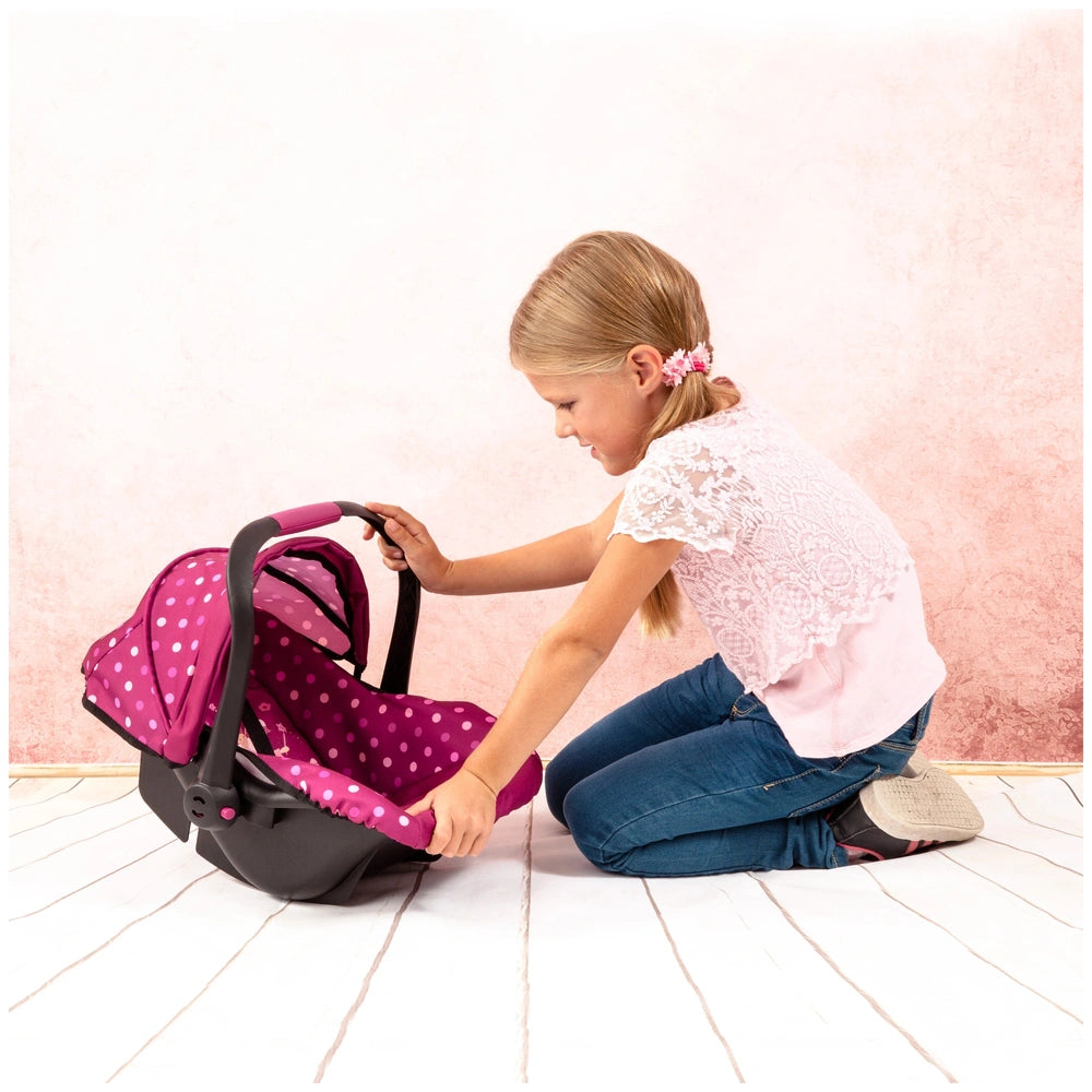 BAYER DESIGN Doll Deluxe Car Seat with Canopy in pink held by a young girl in a white top and blue jeans on light wood floori
