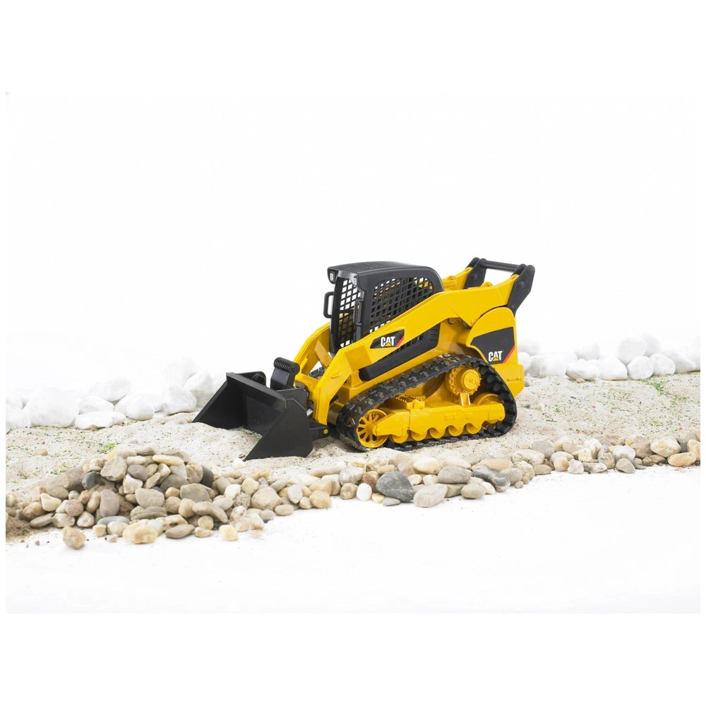 Yellow Bruder CAT Compact Track Loader toy on rocky terrain with rubber tracks and black scoop, for children 3 and up.