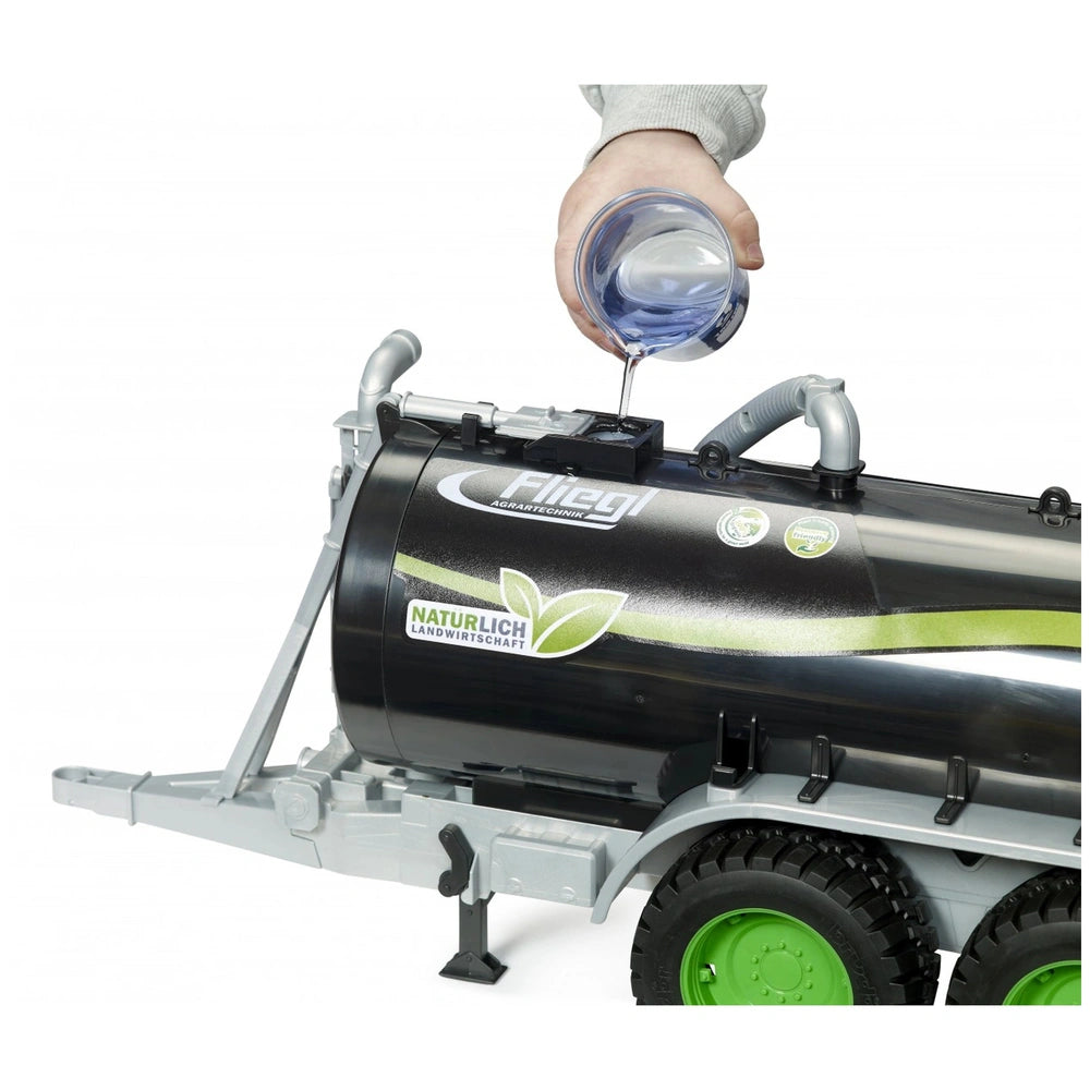 Hand pouring water into the black Bruder Fliegl Barrel Trailer Jumbo Line with green wheels, simulating farming play.