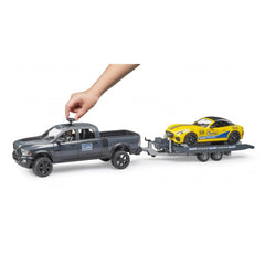 Hand interacting with a gray Bruder RAM Power Wagon toy towing a yellow Roadster on a trailer.