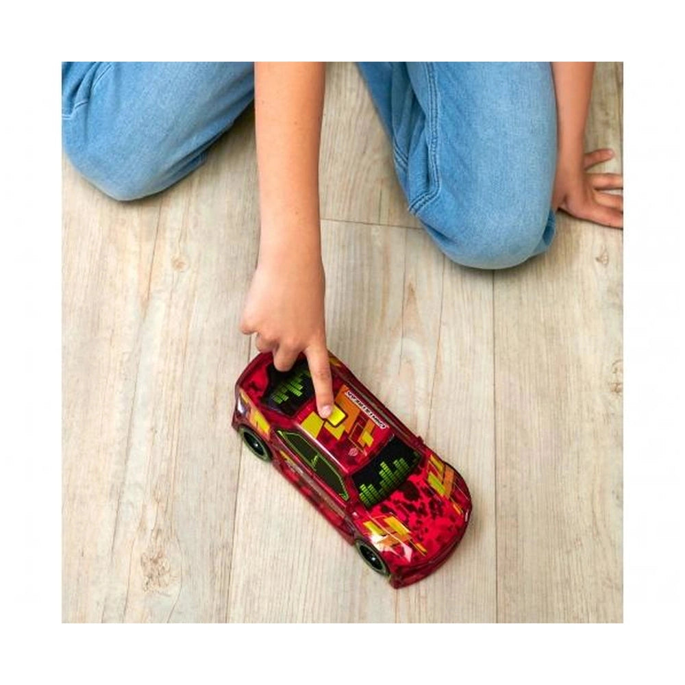 Red toy car with yellow and green details on a light brown wooden floor, with a person's legs in blue jeans in the background