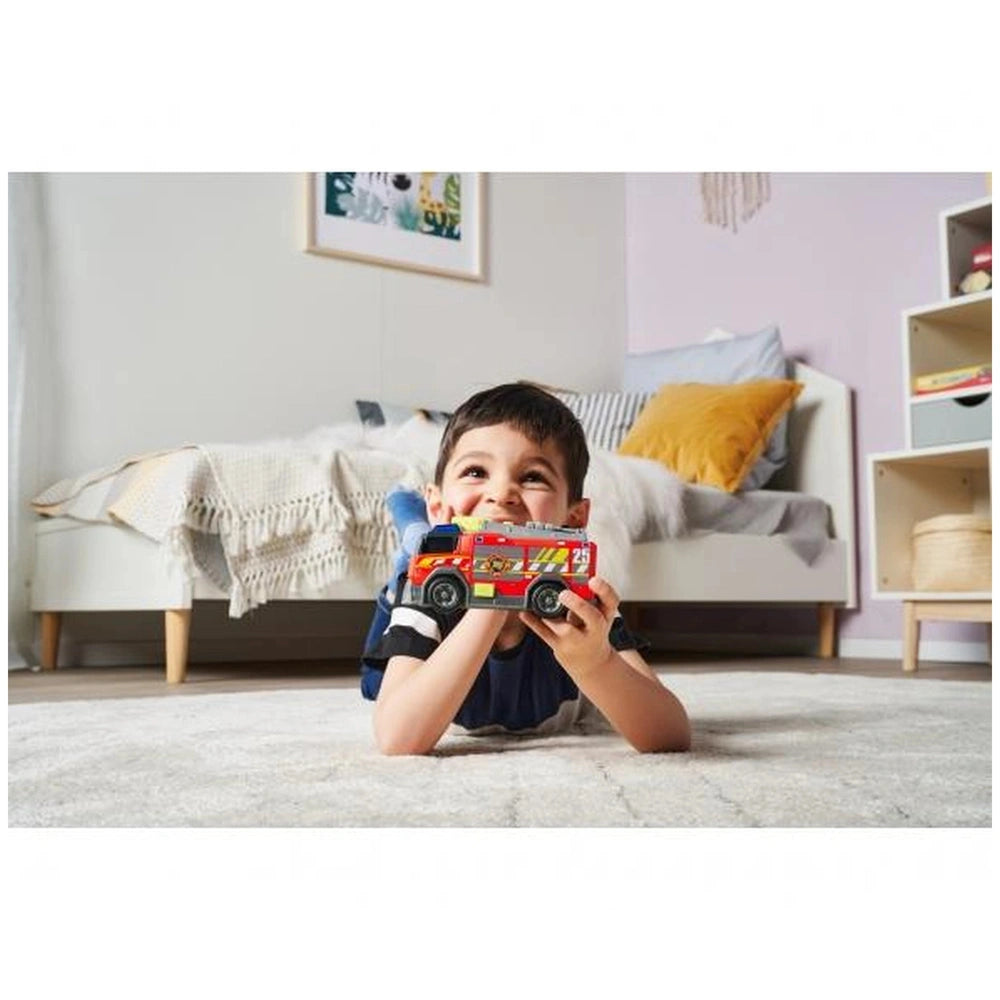 Boy in dark shirt holds red Dickie fire truck with lights and sounds, surrounded by white couch and bed in bright room.