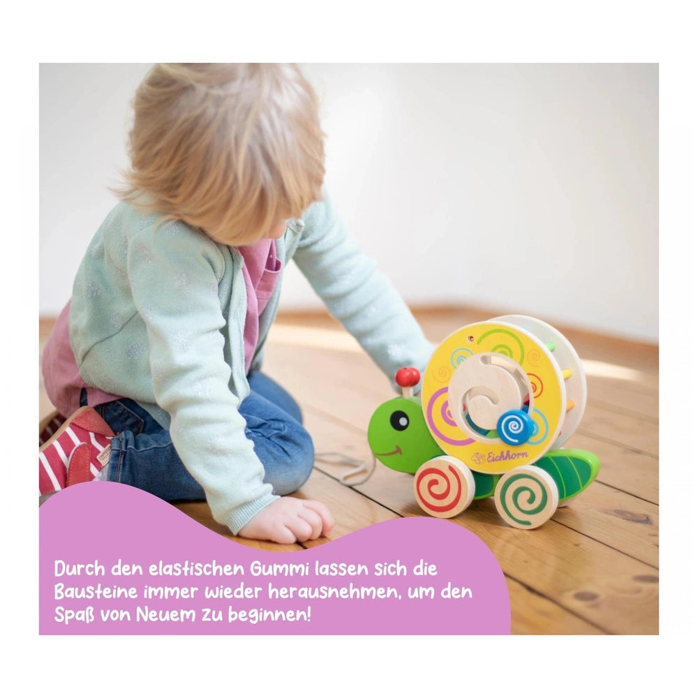 Toddler with blonde hair in a light blue jacket interacts with a green and yellow caterpillar toy on a wooden floor.