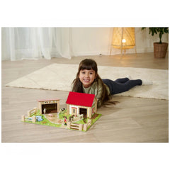 Eichhorn Wooden Farm playset with a smiling girl playing on a rug, featuring a house, cows, sheep, and a cozy indoor setting.