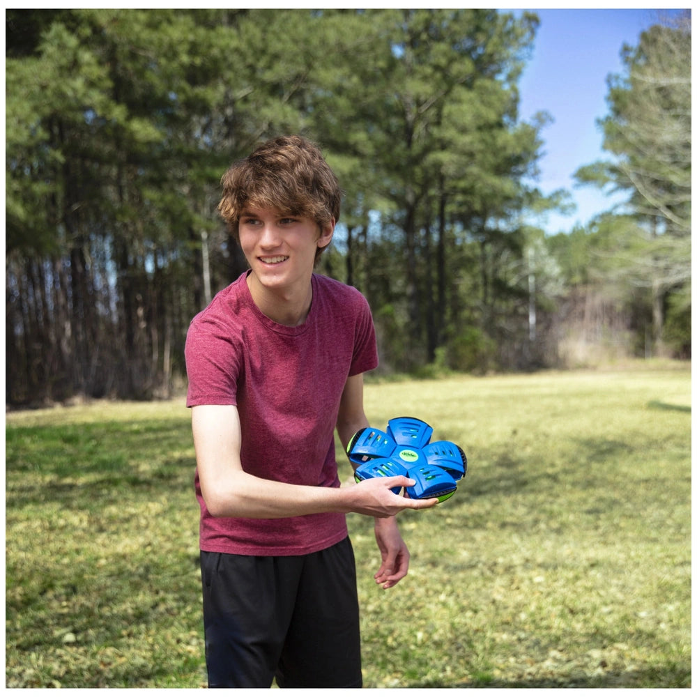 Young man in a red t-shirt holds a blue Wahu Phlat Ball frisbee with green and yellow circles in a green grassy setting.