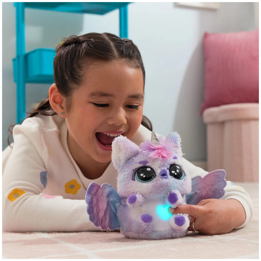 A young girl in a floral top smiles while interacting with a purple and white plush Hatchimals toy that glows blue.