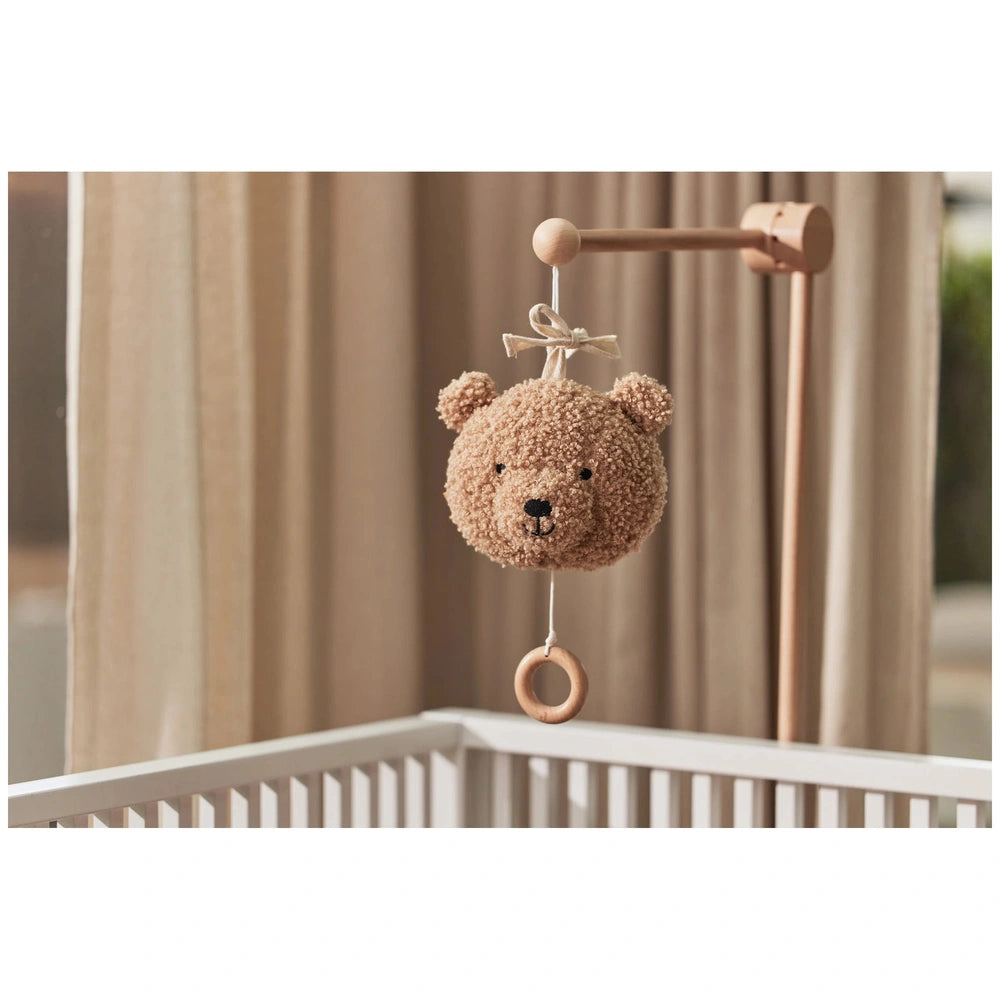 Brown teddy bear stuffed animal hanging from a brown wooden hook on a white railing with a beige curtain in the background.