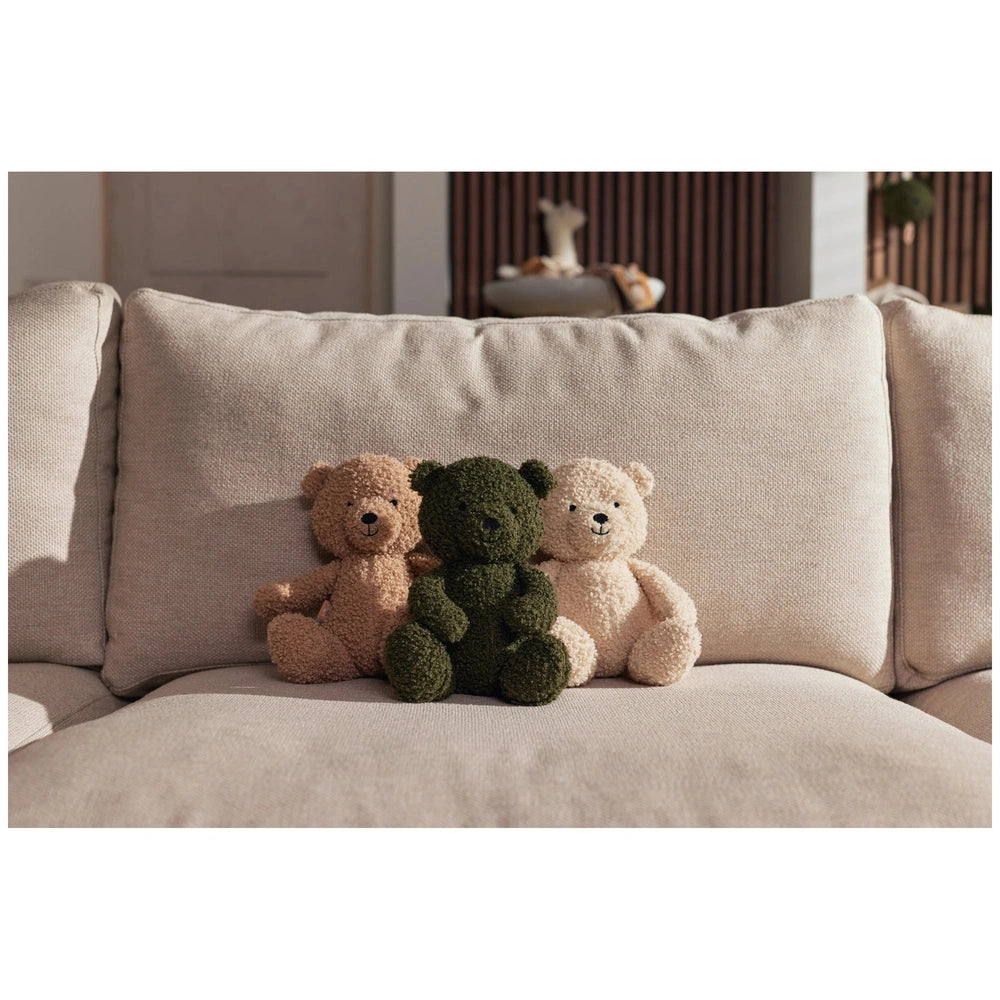 Jollein teddy bears in brown and green sit upright on a light beige couch, surrounded by a softly blurred room background.