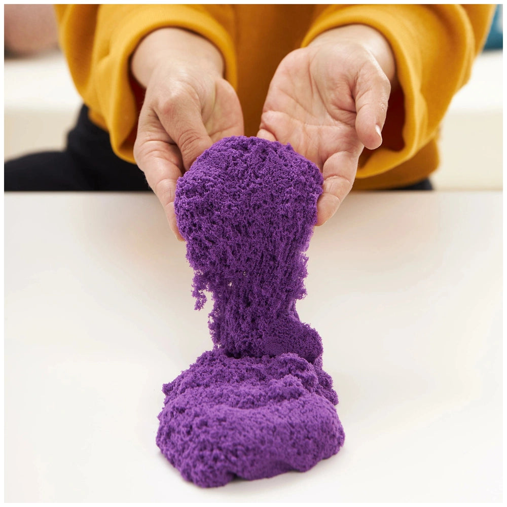 Kinetic Sand Sandbox Set features a person in a yellow sweater holding vibrant purple sand indoors on a white surface.