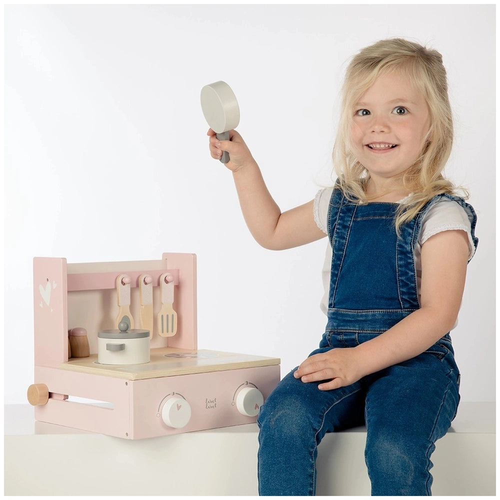 Blonde girl in denim jumpsuit plays with pink and white wooden foldable cooker kitchen set on a white surface.