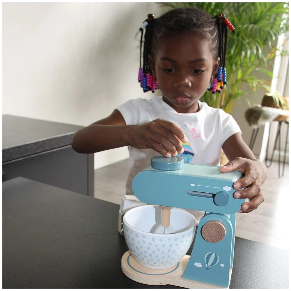 A child in a white shirt and colorful hair accessories plays with a food processor on a dark wood table.