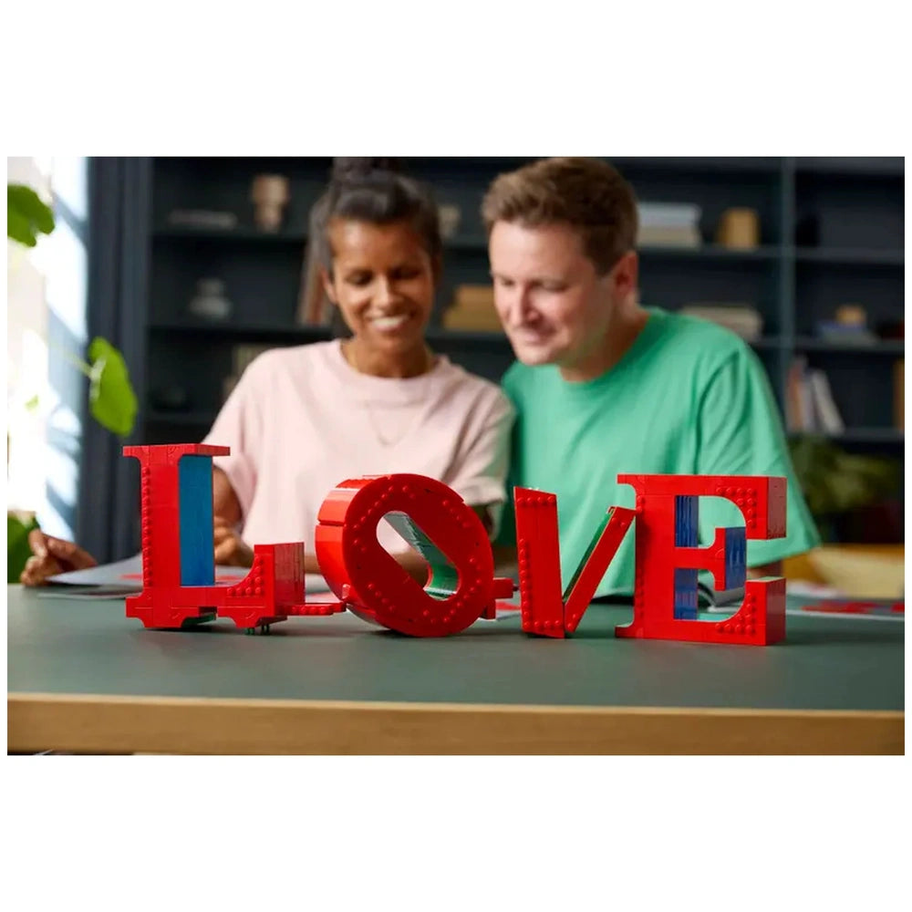 Lego construction set spelling "love" held by a woman, while a smiling man looks on in a cozy bookshelf setting.