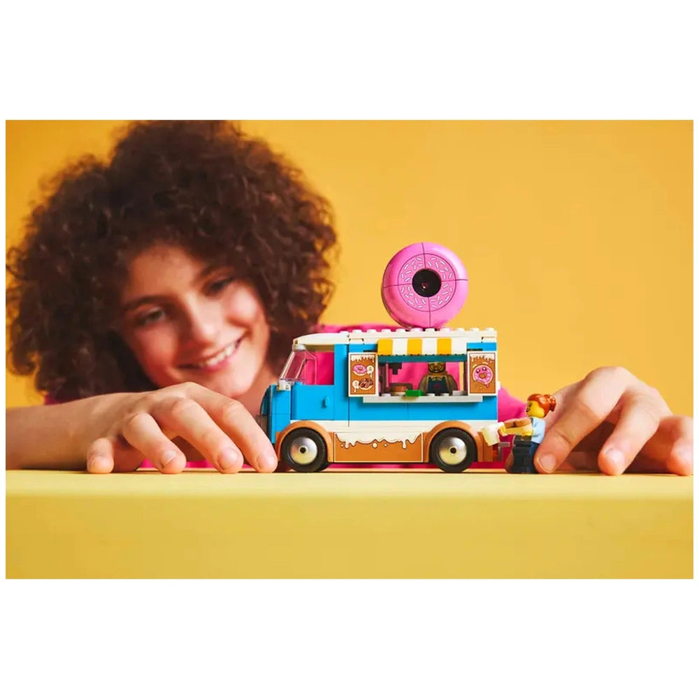 A child smiles while playing with the LEGO City Donut Truck set, featuring a colorful truck with a giant donut on top and a service window. The child is reaching towards the truck, which has a vibrant blue and pink design, showcasing various donut graphics. A mini figure is positioned at the service window, engaging in imaginative play against a bright yellow background.