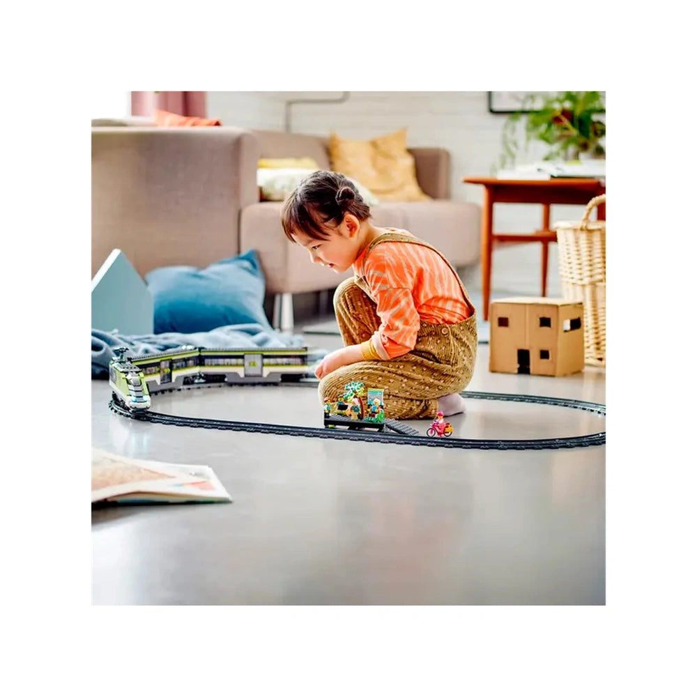 A young child sits on the floor, focused on a colorful LEGO City Express Passenger Train set arranged on a circular track. The child wears an orange-striped shirt and brown overalls, surrounded by plush cushions and furniture in a cozy living room.