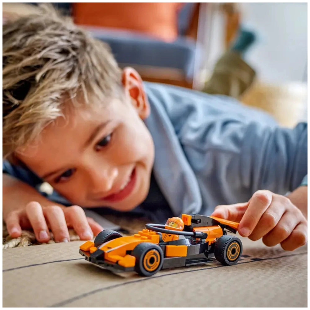 Young boy with short blonde hair excitedly playing with a colorful LEGO® McLaren race car on a cardboard surface.