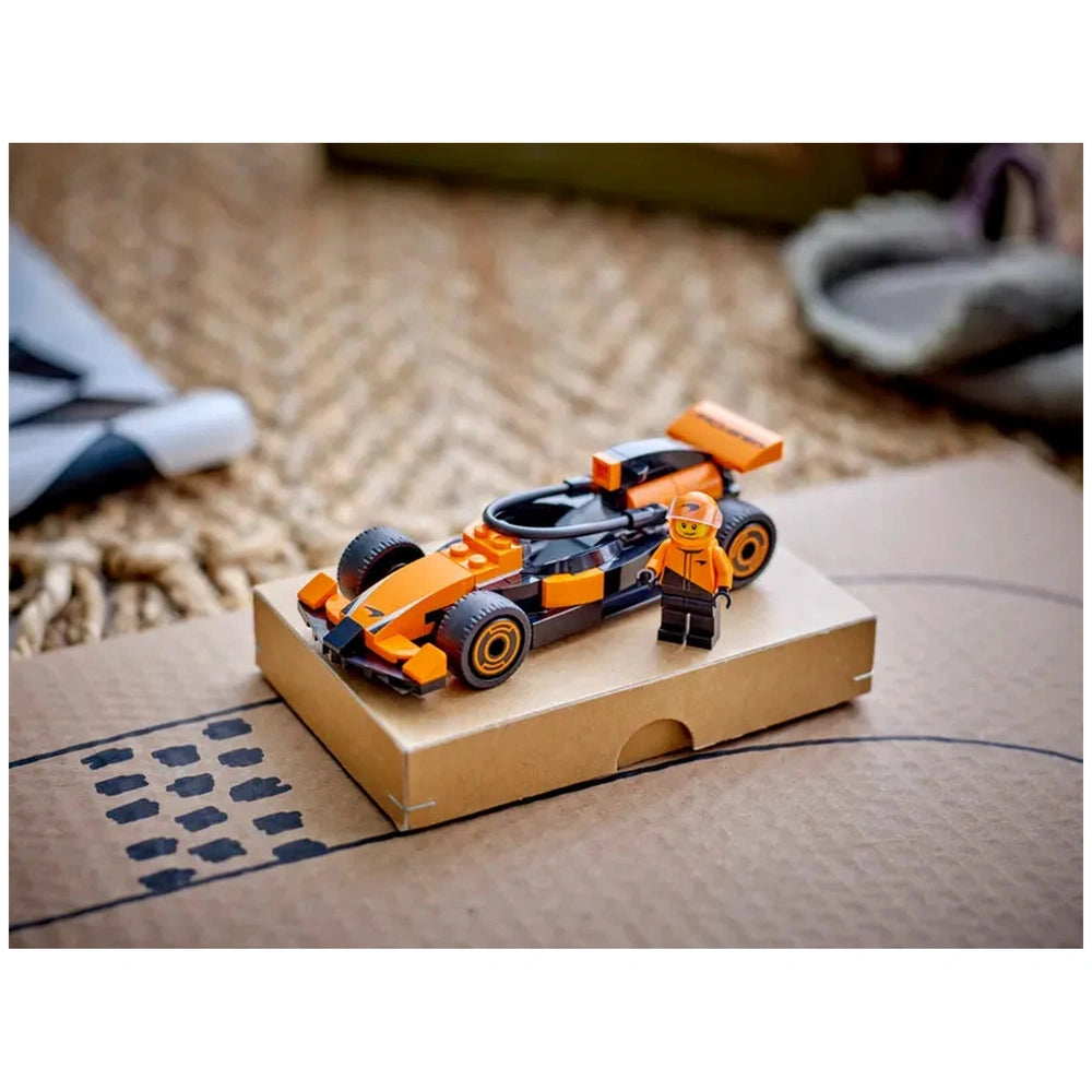 LEGO® City F1® Driver stands next to a detailed McLaren race car on a cardboard platform, featuring a sleek orange and black design.
