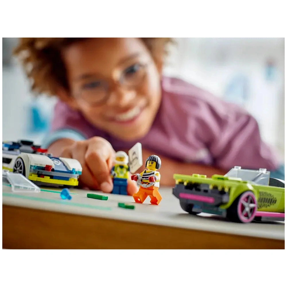 A child in a pink shirt smiles while reaching toward LEGO® figures in a playful scene with a police car and a muscle car on a wooden table.