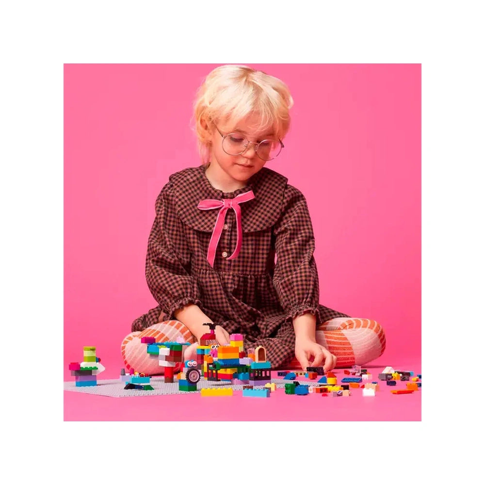 A child with short blond hair, wearing a plaid dress with a pink bow, sits on a pink background, focused on building with colorful LEGO bricks scattered around a gray baseplate.