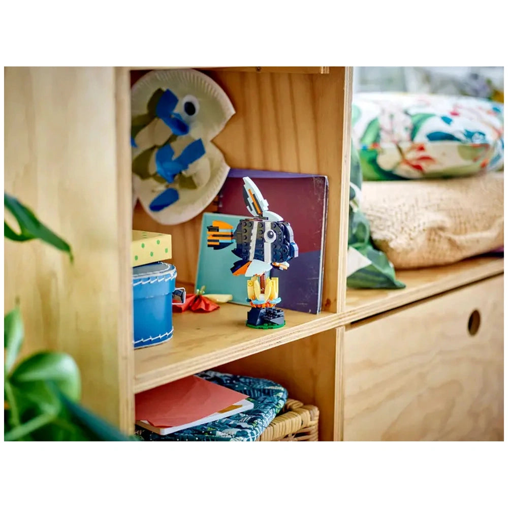 Colorful LEGO Tropical Toucan centerpiece on a wooden shelf, surrounded by books, a blue container, and craft items.