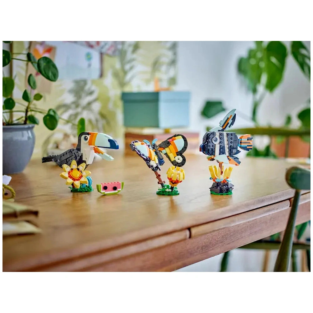 Colorful LEGO creations on a wooden table, featuring a toucan, butterfly, and fish, surrounded by flowers and a slice of watermelon.