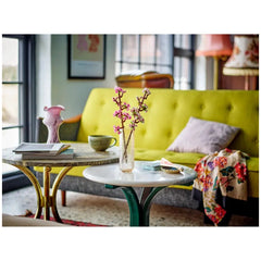 A bright, modern living space featuring a vibrant yellow sofa with a lavender cushion. On two coffee tables, a delicate glass vase holds pink cherry blossom twigs, accompanied by a green mug, magazines, and decorative trinkets. Natural light pours in from large windows, enhancing the cozy atmosphere.