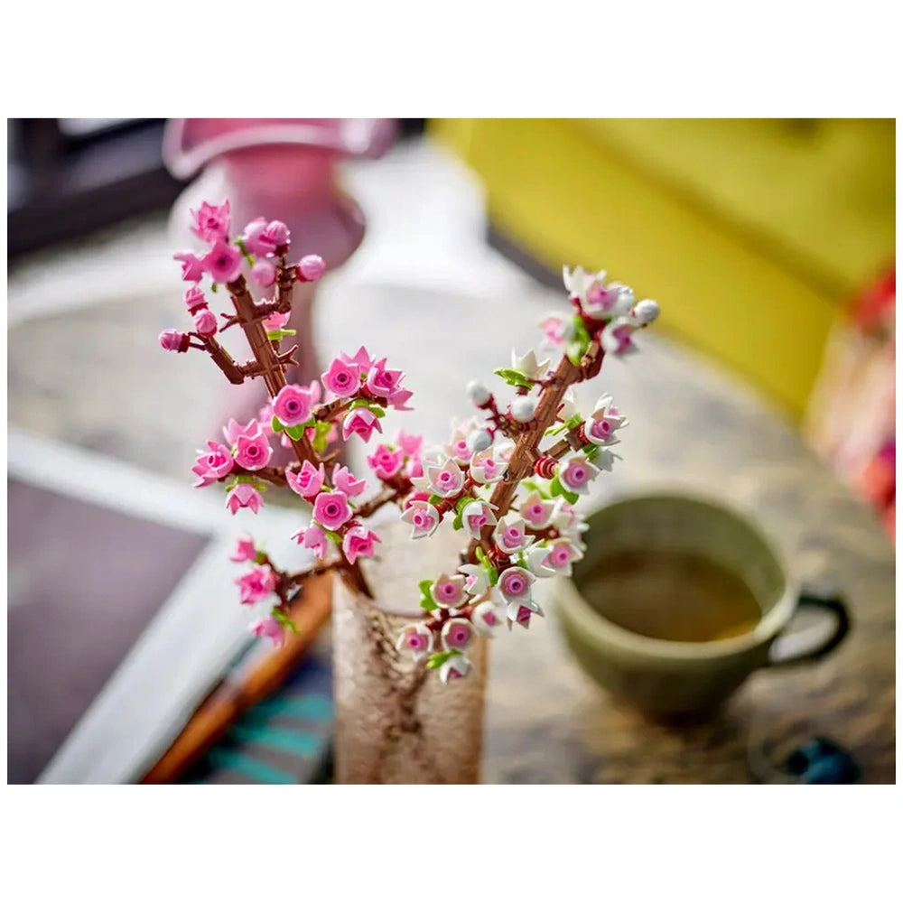 Delicate LEGO® Creator Cherry Blossom twigs in pink and white arranged in a clear vase, with a green cup nearby, set on a textured table.