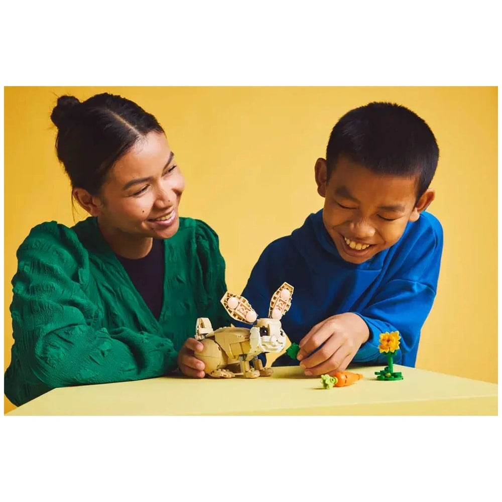 Two children joyfully playing with the LEGO Creator Cute Bunny set on a yellow table, featuring a posable bunny, a carrot, and a sunflower.