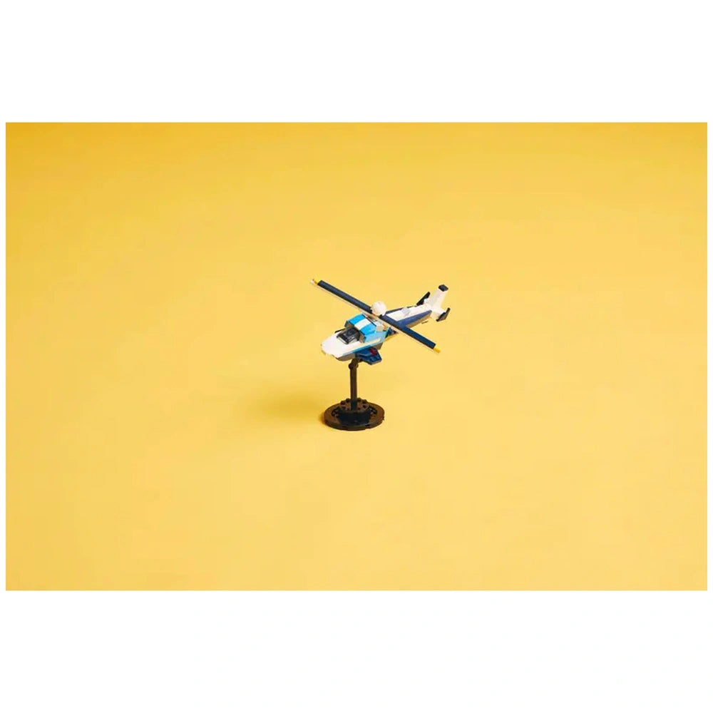 LEGO Creator 3in1 helicopter model displayed on a black stand, featuring a white and blue design with a spinning rotor blade, set against a bright yellow background.
