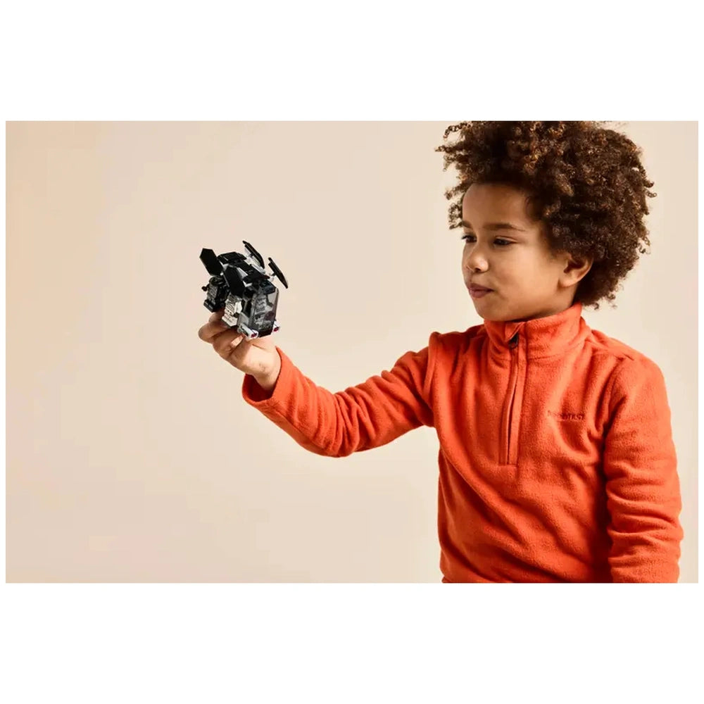 A young boy with curly hair in an orange fleece pullover smiles while holding a LEGO Death Trooper dropship in a playful pose.