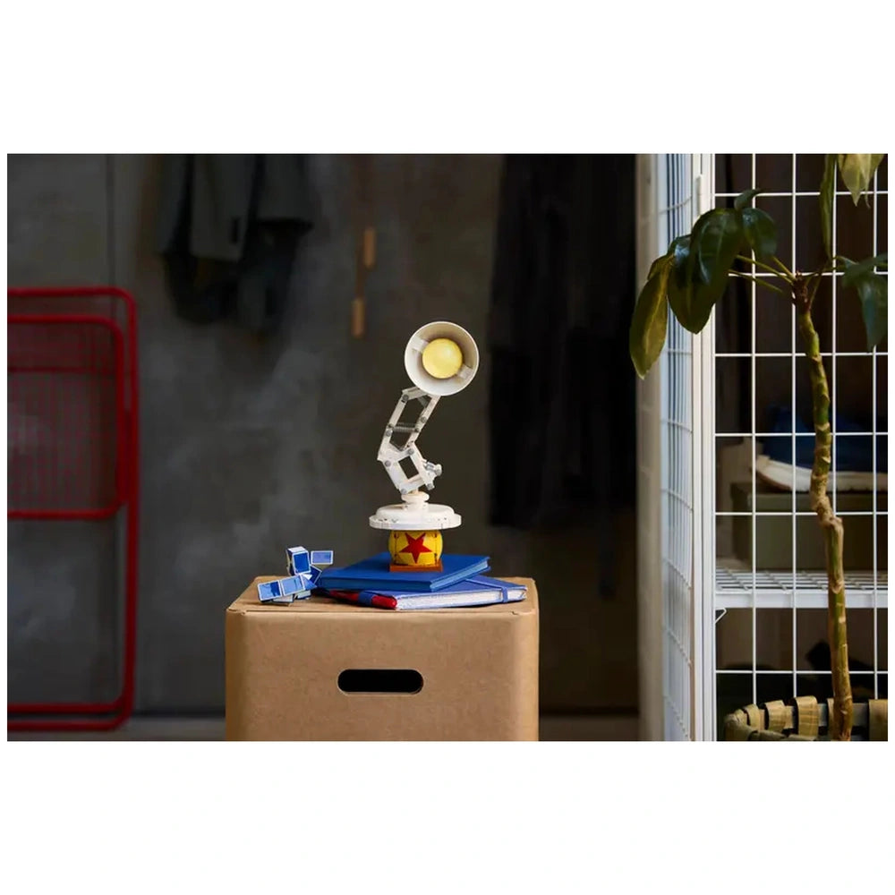 LEGO Disney Pixar Luxo Jr. model displays its iconic lamp design on a stack of blue notebooks atop a cardboard box, with a green plant nearby.