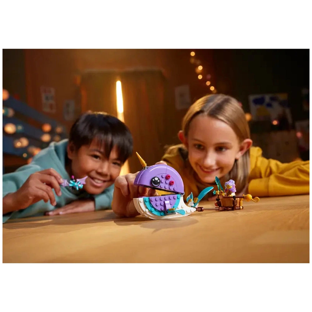 Two children playing with the LEGO® DREAMZzz™ Izzie's Narwhal Hot-Air Balloon set, smiling as they interact with a whimsical narwhal figure and accessories on a wooden table.