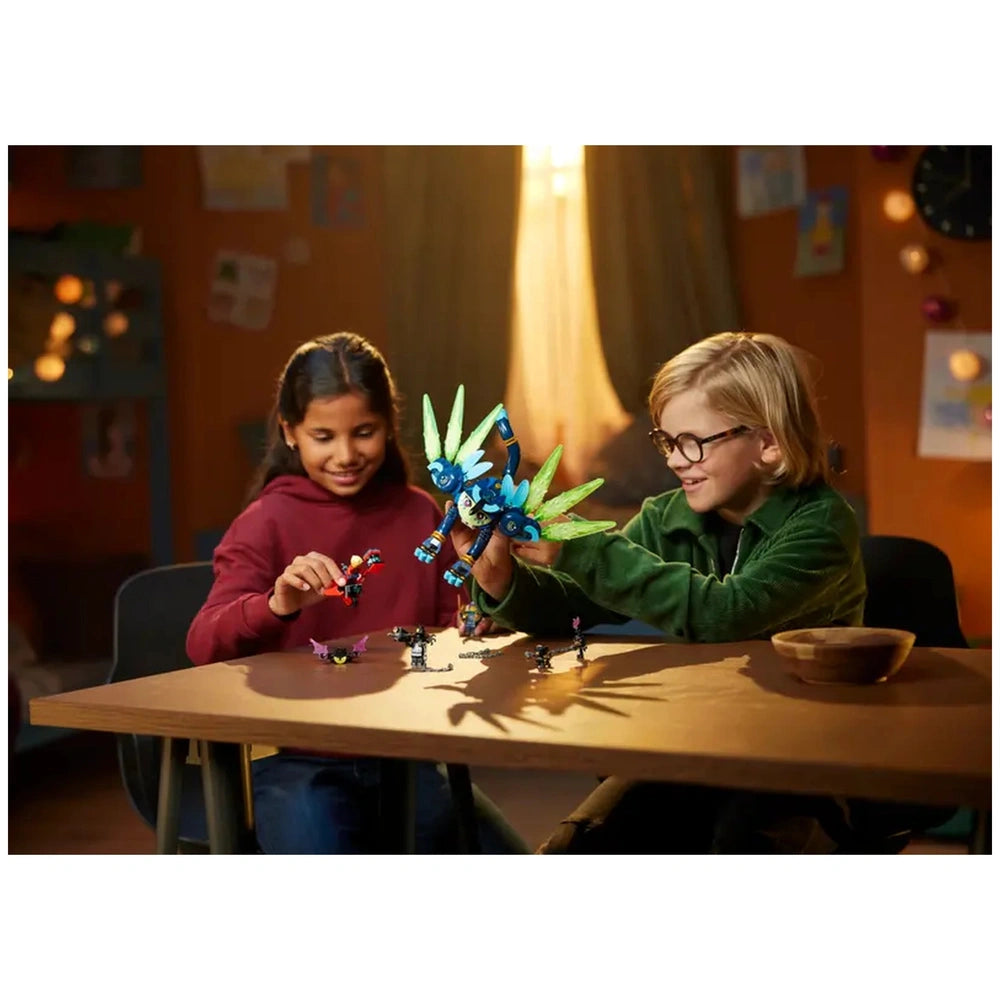 Two children are joyfully playing with LEGO® DREAMZzz™ figures on a wooden table. The girl, wearing a red hoodie, holds a small character, while the boy, in a green sweater, showcases a vibrant blue Cat-Owl figure with green wings. Various LEGO pieces are scattered across the table, and warm light filters through the background, creating a cozy atmosphere.